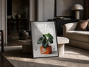 Monstera plant in Nike shoe box print in a black frame leaning on a travertine table in a sunlit lounge