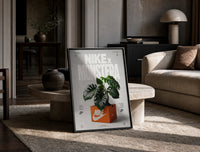 Monstera plant in Nike shoe box print in a black frame leaning on a travertine table in a sunlit lounge