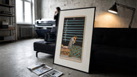 Person seated with nine swords print in a slim black frame leaning on a dark sofa in a concrete print studio