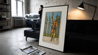 Man holding wooden staff among wands print in a slim black frame leaning on a dark sofa in a concrete print studio