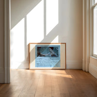 Elegant woman holding hand mirror print in an oak frame on a sunlit wood floor beneath tall window light