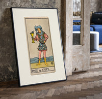 Page holding cup with fish print in a black frame leaning beside a vintage door on parquet