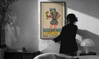 Page of Cups print in a slim black frame being hung on a sunlit wall in a monochrome room