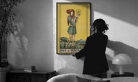 Page of Pentacles print in a slim black frame being hung on a sunlit wall in a monochrome room