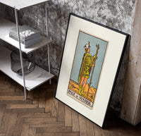 Page of Wands tarot figure poster in a black frame leaning beside a shelf in a raw-plaster loft