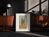Black-framed Page of Wands tarot figure print on a moody console with designer coffee-table books