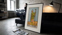 Page of Wands tarot figure print in a slim black frame leaning on a dark sofa in a concrete print studio