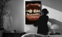 Parted Lips with Gold-Tone Grillz print in a slim black frame being hung on a sunlit wall in a monochrome room