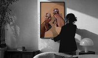 Cowhide Mules and Chain Anklet print in a slim black frame being hung on a sunlit wall in a monochrome room