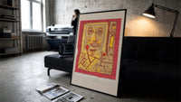 Abstract face with small figures print in a slim black frame leaning on a dark sofa in a concrete print studio