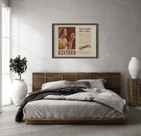 Woman holding hand mirror print in a dark wood frame above a rattan bed on a plaster wall
