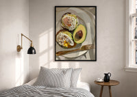 Poached Eggs on Avocado Toast print in a black frame above a bed with striped linens, a brass wall lamp and a sunlit window