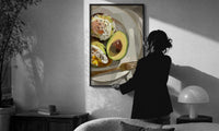 Poached Eggs on Avocado Toast print in a slim black frame being hung on a sunlit wall in a monochrome room