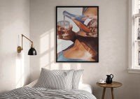 Pouring a Sparkling Drink print in a black frame above a bed with striped linens, a brass wall lamp and a sunlit window