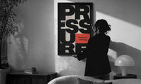 Pressure Is a Privilege print in a slim black frame being hung on a sunlit wall in a monochrome room