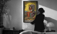 Queen of Pentacles print in a slim black frame being hung on a sunlit wall in a monochrome room