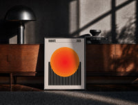 Black-framed Radiate 2026 Orange Sun print on the floor in front of a dark wooden console
