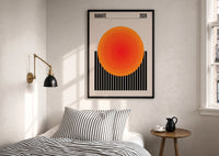 Radiate 2026 Orange Sun print in a black frame above a bed with striped linens, a brass wall lamp and a sunlit window