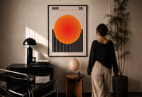 Radiate 2026 Orange Sun print in a black frame on a sunlit plaster wall, a woman looking on beside a mushroom lamp