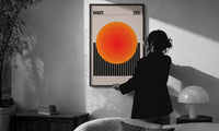 Radiate 2026 Orange Sun print in a slim black frame being hung on a sunlit wall in a monochrome room