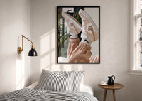 Raised Legs in Beige Sneakers print in a black frame above a bed with striped linens, a brass wall lamp and a sunlit window