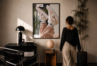 Raised Legs in Beige Sneakers print in a black frame on a sunlit plaster wall, a woman looking on beside a mushroom lamp