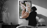 Raised Legs in Beige Sneakers print in a slim black frame being hung on a sunlit wall in a monochrome room