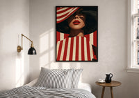 Red Stripes and a Wide Hat print in a black frame above a bed with striped linens, a brass wall lamp and a sunlit window