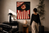 Red Stripes and a Wide Hat print in a black frame on a sunlit plaster wall, a woman looking on beside a mushroom lamp