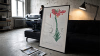 Botanical illustration of pink lily print in a slim black frame leaning on a dark sofa in a concrete print studio
