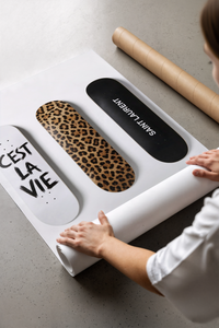 Hands unrolling Saint Laurent Cest La Vie Skateboards mockup displaying leopard print and black designs on white.