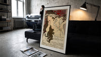 Woman with flowing hair holding feather print in a slim black frame leaning on a dark sofa in a concrete print studio