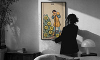 Seven of Pentacles print in a slim black frame being hung on a sunlit wall in a monochrome room