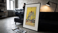 Man carrying five swords print in a slim black frame leaning on a dark sofa in a concrete print studio