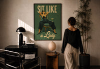 Sit Like a Lady print in a black frame on a sunlit plaster wall, a woman looking on beside a mushroom lamp