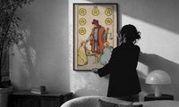 Six of Pentacles print in a slim black frame being hung on a sunlit wall in a monochrome room