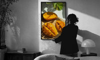 Sliced Mango print in a slim black frame being hung on a sunlit wall in a monochrome room