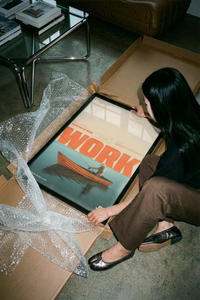 Framed orange WORK text poster showing a man fishing from a wooden boat on a wall