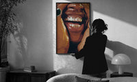 Framed Grillz Smile print in a slim black frame being hung on a sunlit wall in a monochrome room