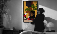 Sneakers and a Plant-Filled Tote print in a slim black frame being hung on a sunlit wall in a monochrome room
