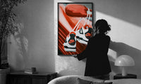Sneakers Hanging from a Car Mirror print in a slim black frame being hung on a sunlit wall in a monochrome room