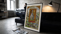 Alchemical crowned flask with child dragon print in a slim black frame leaning on a dark sofa in a concrete print studio