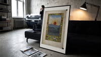 Sun with human face over town print in a slim black frame leaning on a dark sofa in a concrete print studio