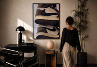 Stacked Brown and Cream Sneakers print in a black frame on a sunlit plaster wall, a woman looking on beside a mushroom lamp