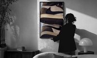 Stacked Brown and Cream Sneakers print in a slim black frame being hung on a sunlit wall in a monochrome room