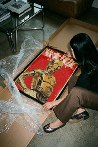 Woman lifting a framed C-3PO Japanese retro print out of a cardboard box with bubble wrap on a concrete floor