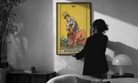 Strength print in a slim black frame being hung on a sunlit wall in a monochrome room