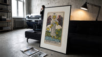 Winged angel pouring water between cups print in a slim black frame leaning on a dark sofa in a concrete print studio