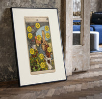 Ten of Pentacles tarot card print in a black frame leaning beside a vintage door on parquet