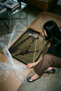 Woman lifting the overhead serve tennis artwork out of a cardboard delivery box near a wall
